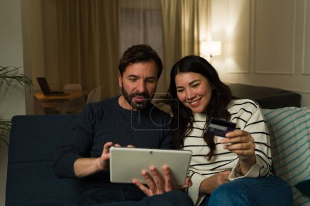 Téléchargez les photos : Attrayant couple marié achats en ligne payer avec une carte de crédit et sourire en utilisant la tablette relaxant sur le canapé - en image libre de droit