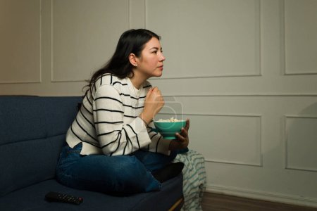 Foto de Hermosa mujer relajada sentada en el sofá de la sala comiendo palomitas de maíz y viendo películas por la noche - Imagen libre de derechos