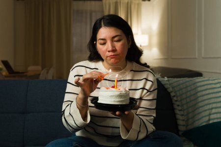 Foto de Triste mujer caucásica solitaria celebrando su cumpleaños sola en casa mientras enciende velas en un pastel sintiéndose deprimida - Imagen libre de derechos