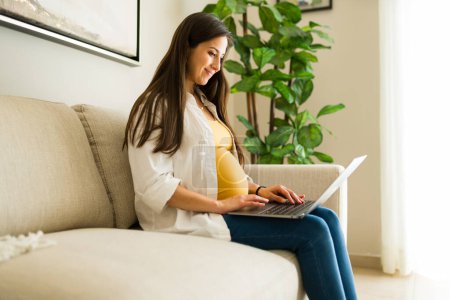 Téléchargez les photos : Belle femme enceinte assise sur le canapé tapant sur l'ordinateur portable et souriant tout en se relaxant dans le salon à la recherche en ligne sur la maternité - en image libre de droit