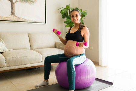 Téléchargez les photos : Magnifique femme enceinte avec un gros ventre souriant tout en soulevant haltère poids exercice avec une balle de fitness dans le salon - en image libre de droit