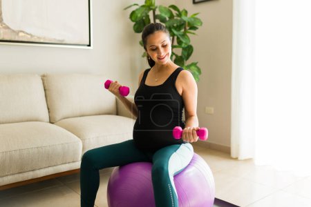 Photo for Caucasian attractive woman with a belly exercising using a fitness ball and dumbbell weights for a healthy maternity and pregnancy - Royalty Free Image