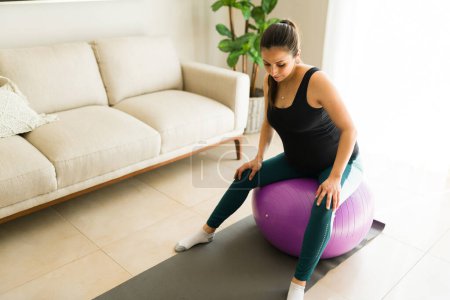 Téléchargez les photos : Fitness femme enceinte faisant une séance d'entraînement à la maison en utilisant une balle de fitness et haltères de levage rester en bonne santé pour l'accouchement et la grossesse - en image libre de droit