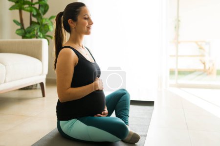 Téléchargez les photos : Heureuse femme enceinte caucasienne touchant son ventre et souriant tout en faisant du yoga à la maison pour une séance d'entraînement saine et active - en image libre de droit