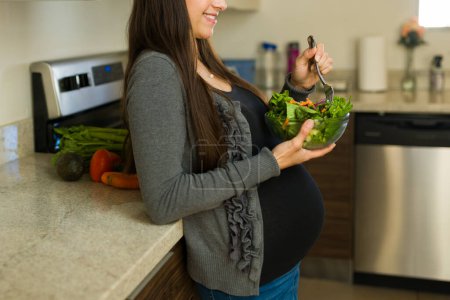Beautiful expectant mother and pregnant woman eating a green salad in the kitchen and smiling while having a healthy pregnancy 