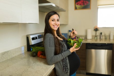 Wunderschöne gesunde schwangere Frau lächelt, während sie einen grünen Salat in der Küche zu Hause isst, während sie eine schöne Schwangerschaft hat