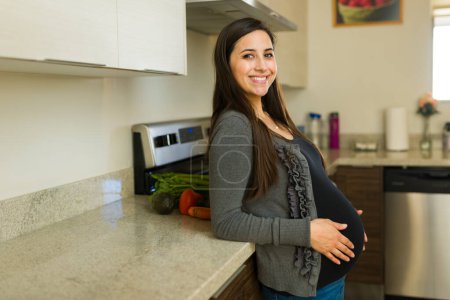 Gorgeous pregnant woman smiling making eye contact while in the kitchen at home thinking about her baby and maternity