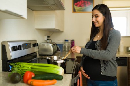 Fröhliche Schwangere berührt ihren schönen Bauch und lächelt, während sie in der Küche gesundes Mittagessen kocht und Gemüsesuppe zubereitet