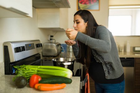 Kaukasische Schwangere schaut hungrig und probiert eine Gemüsesuppe für eine gesunde Schwangerschaft, während sie in der Küche kocht