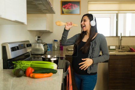 Aufgeregte Schwangere, die mit Kopfhörern Musik hört, während sie in der Küche ein gesundes Mittagessen zubereitet und ihre Schwangerschaft genießt