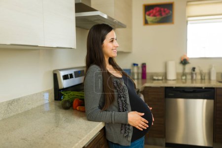 Fröhliche schwangere Frau lächelt, während sie in der Küche bereit zum Mittagessen ist, und sieht glücklich aus, als sie ihren Bauch berührt und die Mutterschaft genießt 