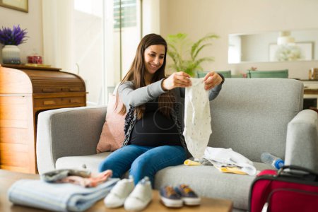Cheerful maternal woman smiling while folding baby clothes while expecting her baby feeling happy about maternity
