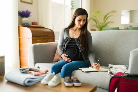 Schöne schwangere Frau, die für ihr zukünftiges Baby und die Geburt plant, während sie ihre Agenda schreibt, während sie Babykleidung faltet
