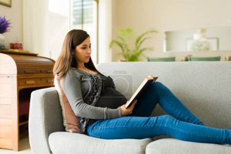 Beautiful pregnant woman reading a book about motherhood and childhood while expecting a baby and resting on the couch
