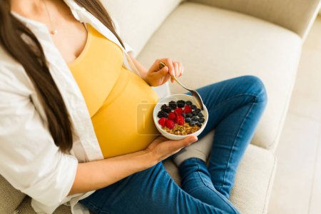 Photo for Top view of a healthy pregnant woman sitting on the couch while eating berries yogurt and healthy food during her pregnancy - Royalty Free Image