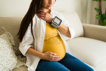 Foto de Caucásica embarazada madre chequeando su presión arterial alta con un monitor mientras muestra su hermoso vientre sentado en la sala de estar - Imagen libre de derechos