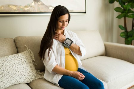 Téléchargez les photos : Femme enceinte en bonne santé assise sur le canapé souriant en utilisant un tensiomètre pour vérifier sa santé pendant sa grossesse - en image libre de droit