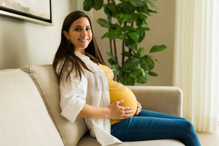 Téléchargez les photos : Femme enceinte souriante faisant un contact visuel tout en se relaxant à la maison se sentant heureuse d'attendre son premier bébé né - en image libre de droit