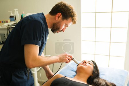 Foto de Terapeuta en la clínica de salud dando una radiofrecuencia electromédica tecar a una joven hispana para un tratamiento de belleza - Imagen libre de derechos