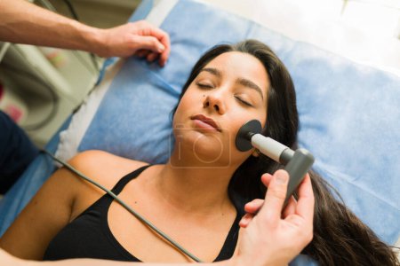 Téléchargez les photos : Vue du dessus d'une femme latine se relaxant tout en obtenant un traitement diathermie ou radiofréquence sur son visage à la clinique de beauté - en image libre de droit