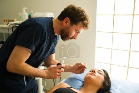 Foto de Terapeuta médico guapo poniendo crema hidratante mientras trata a una mujer para un tratamiento de belleza de radiofrecuencia en la clínica de salud - Imagen libre de derechos