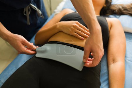 Acercamiento de una mujer joven que recibe terapia de hipertermia en su abdomen mientras habla con su médico en la clínica