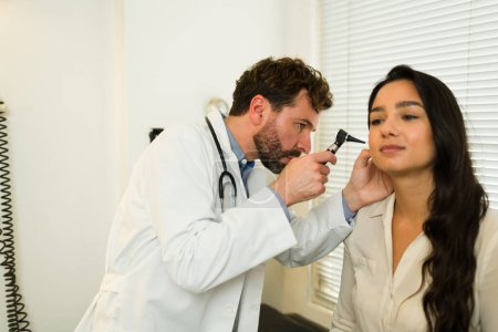 Atractivo médico durante una consulta de chequeo con un otoscopio mientras revisa la oreja de una hermosa paciente femenina 