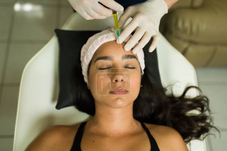 Téléchargez les photos : Angle élevé d'une femme hispanique attrayante à la clinique de santé recevant des injections de plasma sur son visage pour un traitement alternatif régénératif - en image libre de droit