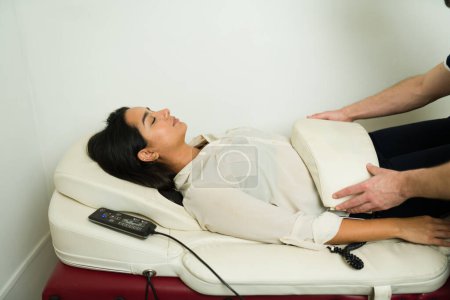 Foto de Mujer latina en la clínica de bienestar recibiendo un masaje de vibración o terapia de andulación utilizando la máquina de curación - Imagen libre de derechos