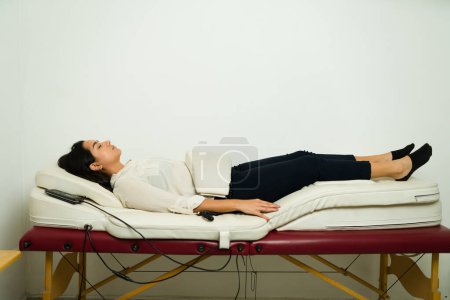 Foto de Mujer latina relajada usando medicina alternativa recibiendo un masaje de terapia de andulación usando vibración en su abdomen para aliviar el dolor - Imagen libre de derechos