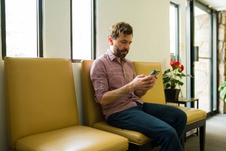 Téléchargez les photos : Latin homme textos sur son smartphone tandis que dans la salle d'attente à la clinique de santé se préparer pour un examen médical avec son médecin - en image libre de droit