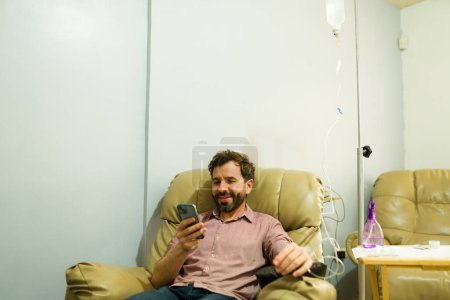 Aufgeregter Mann in der Klinik lächelt, während er IV-Therapie und Vitamincocktail bekommt, um das System wiederherzustellen, während er auf dem Smartphone SMS schreibt