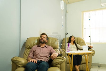 Hispanic couple getting alternative medicine at the health clinic with an IV drip with myers vitamin cocktail looking happy and relaxed