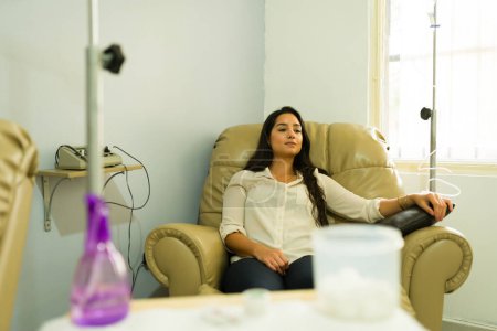 Beautiful latin woman relaxing and smiling while getting an IV drip and vitamin therapy at the alternative medicine clinic