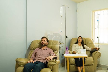 Foto de Mujer y hombre hispanos en sus 30 años en la clínica de salud recibiendo suero vitaminizado con un tratamiento intravenoso por goteo - Imagen libre de derechos