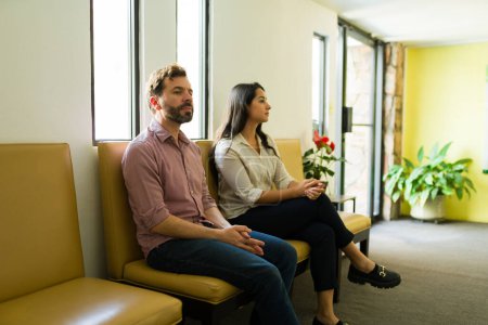 Téléchargez les photos : Couple latin assis ensemble dans la salle d'attente de la clinique de bien-être et attendant le médecin ou le thérapeute - en image libre de droit
