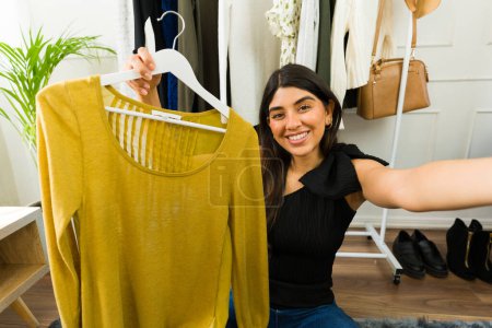 Foto de Mujer joven y feliz sostiene un suéter amarillo, seleccionando trajes en su acogedor dormitorio - Imagen libre de derechos
