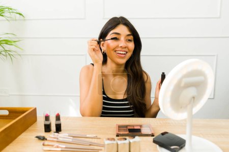 Photo for Cheerful young woman enjoys a beauty routine, holding mascara at a well-organized makeup table - Royalty Free Image