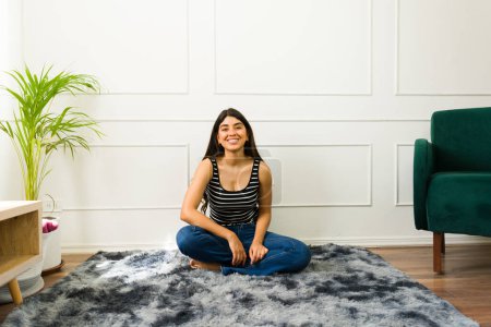 Téléchargez les photos : Femme hispanique souriante assise les jambes croisées sur un tapis dans un salon lumineux - en image libre de droit