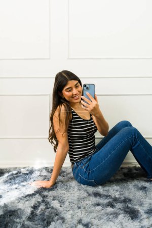 Fröhliche junge Frau in lässiger Kleidung macht ein Selfie, während sie drinnen auf einem flauschigen Teppich sitzt
