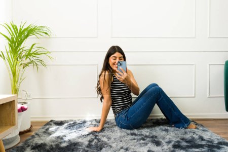 Téléchargez les photos : Joyeux jeune femme assise sur un tapis et capturant un selfie avec son téléphone - en image libre de droit