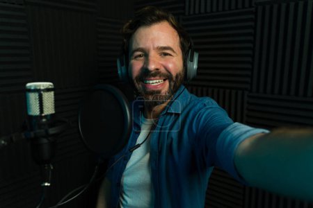 Hombre alegre con auriculares tomando una selfie mientras graba en un estudio de podcast insonorizado