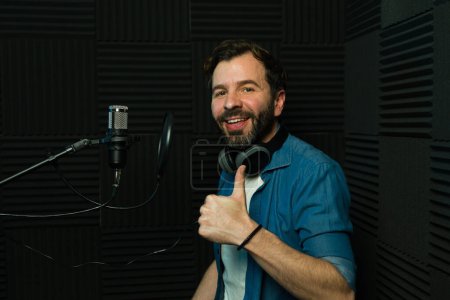 Foto de Hombre alegre con auriculares posa en un estudio insonorizado, haciendo gestos de aprobación durante una sesión de música - Imagen libre de derechos