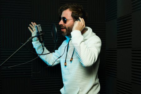 Téléchargez les photos : Homme barbu avec des lunettes de soleil chante passionnément dans un microphone, portant des écouteurs dans un studio d'enregistrement insonorisé - en image libre de droit