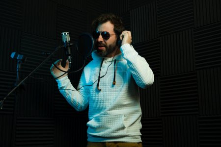 Foto de Hombre barbudo con auriculares canta apasionadamente en un micrófono con espuma de estudio oscuro en el fondo - Imagen libre de derechos