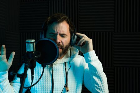 Téléchargez les photos : Chanteuse passionnée avec écouteurs se produit dans une cabine d'enregistrement insonorisée, éclairée par la lumière bleue - en image libre de droit
