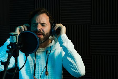 Téléchargez les photos : Chanteur masculin avec écouteurs performant en studio d'enregistrement insonorisé, expression artistique - en image libre de droit