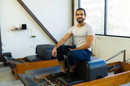 Photo for Cheerful male practitioner sitting on a pilates reformer bed in a well-lit exercise room - Royalty Free Image