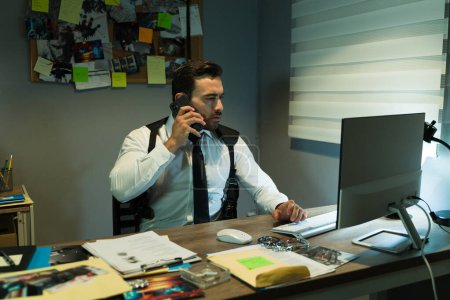 Foto de Investigador de la policía caucásica investigando un caso en su computadora en una oficina con poca luz y hablando por teléfono - Imagen libre de derechos