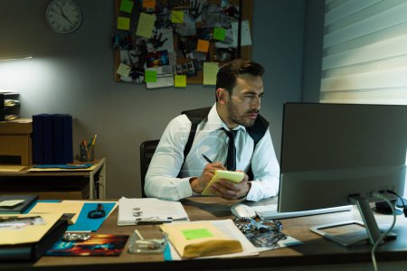 Foto de Detective reuniendo evidencia en una oficina débilmente iluminada rodeada de archivos de casos y un escritorio abarrotado mirando la pantalla de una computadora - Imagen libre de derechos